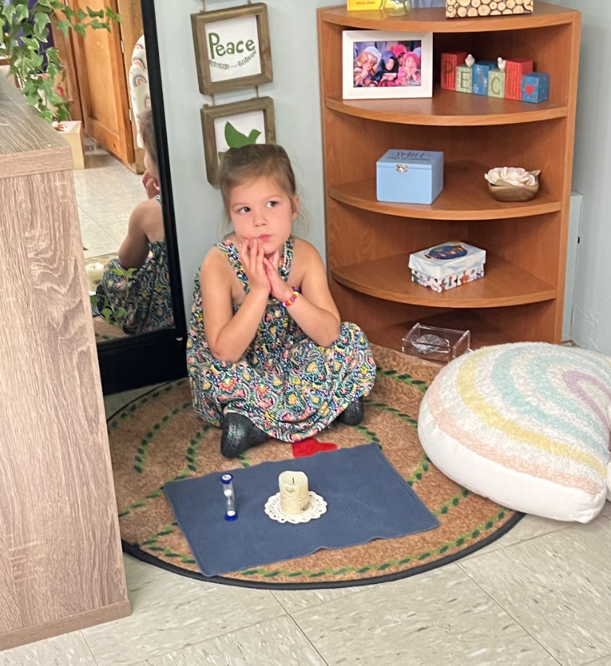 A young student sitting cross-legged on a labyrinth rug in a classroom setting, with a small labyrinth rug and decorative items in the background.