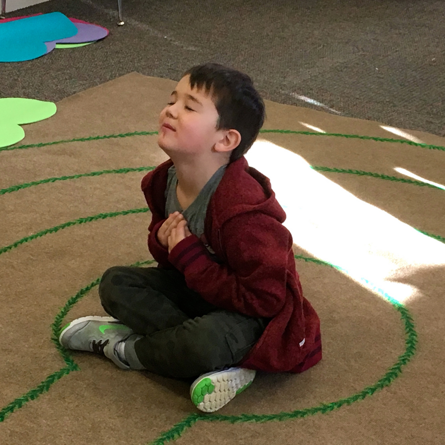 A child sitting cross-legged on the floor with eyes closed, appearing to be in a state of relaxation or meditation, with a labyrinth pattern drawn on the floor in front of him.